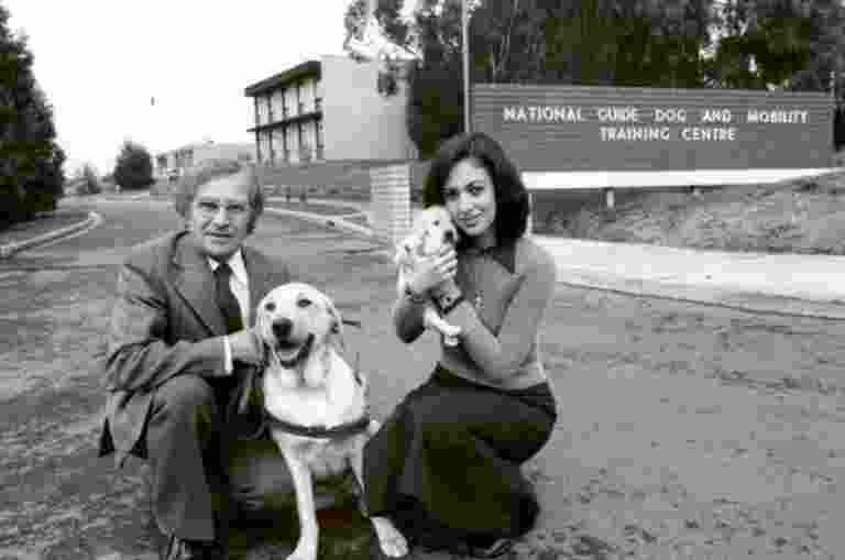 A 1940's black and white image of two people outside a Guide Dog Training Centre. One of the people in kneeling down next to a yellow labrador Guide Dog in harness and the other is holding a yellow ten week old labrador puppy. Both dogs and people are looking at the camera.