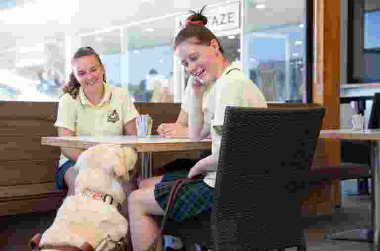 Two young adults sitting a cafe table. A yellow Guide Dog in harness is seated next to the table looking up at the people. The young adults are looking down at the Guide Dog smiling.