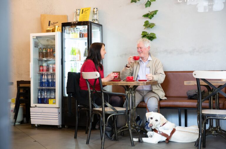 Two people seated at a cafe table looking at each smiling. Next to the table, at the feet of one of the people at the table, is a yellow labrador Guide Dog in harness who is looking at the camera.