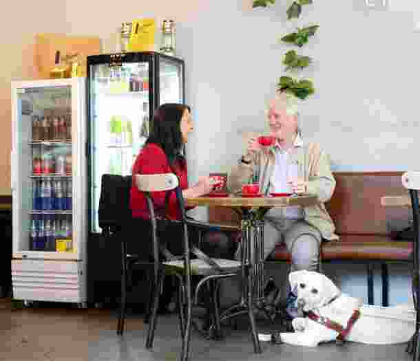 Two people seated at a cafe table looking at each smiling. Next to the table, at the feet of one of the people at the table, is a yellow labrador Guide Dog in harness who is looking at the camera.