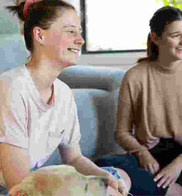 Three young adults sitting inside with a yellow labrador dog next to one of them. They are all looking at each other smiling.