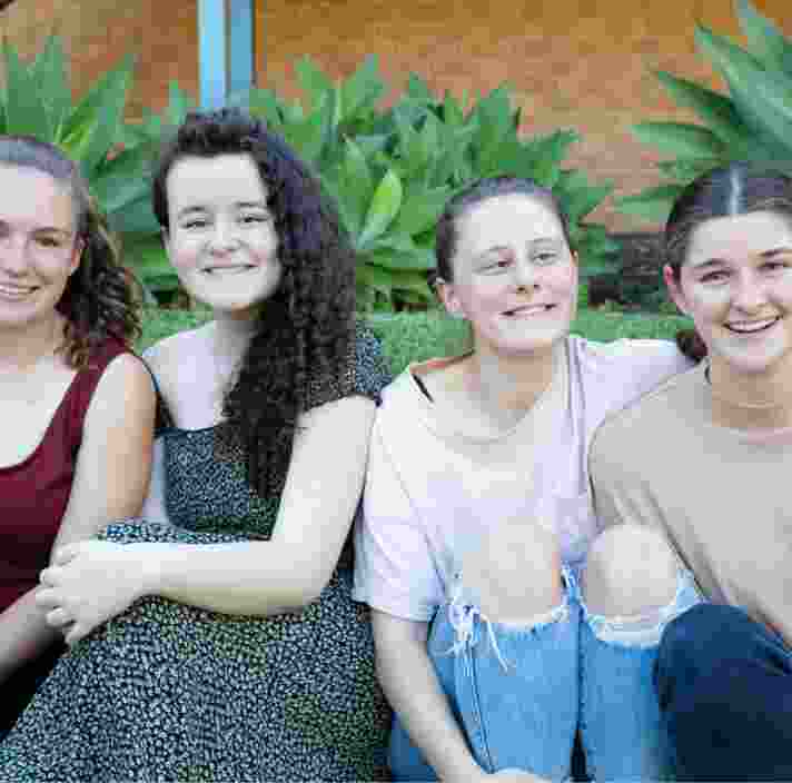A group of four young adults sitting outside on the grass all in a row. Everyone is looking at the camera smiling.