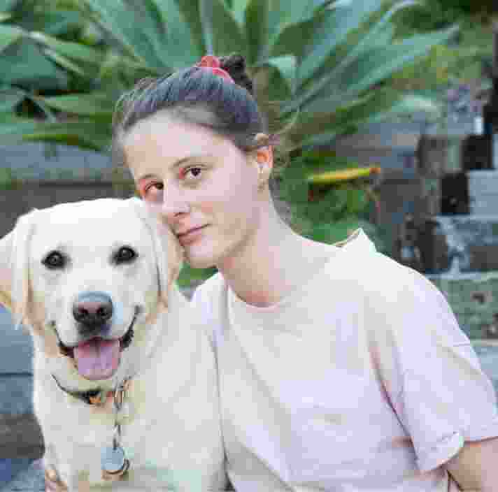 A person looking straight at the camera with their arm around a yellow labrador dog. They are seated at the bottom of a set of stairs.