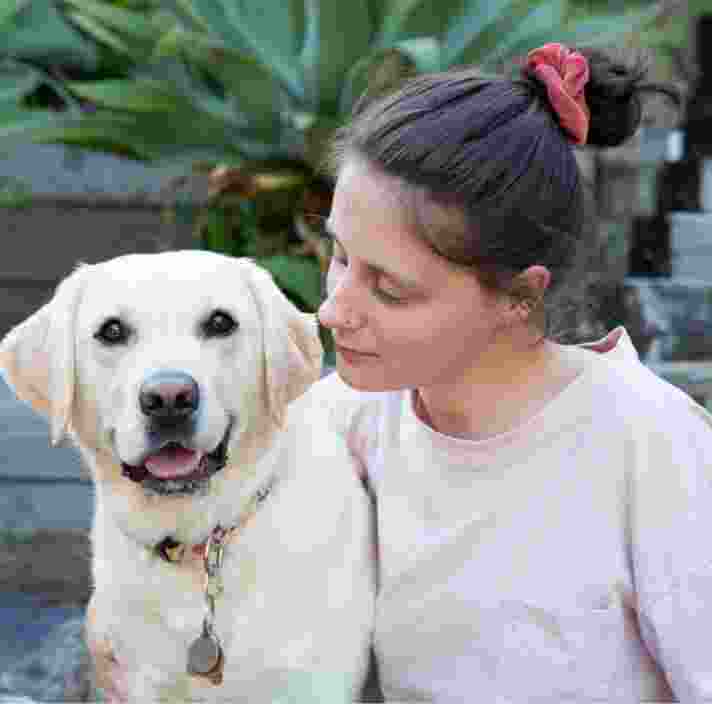 A person seated next to a yellow labrador dog, The person is looking at the dog and the dog is looking at the camera.