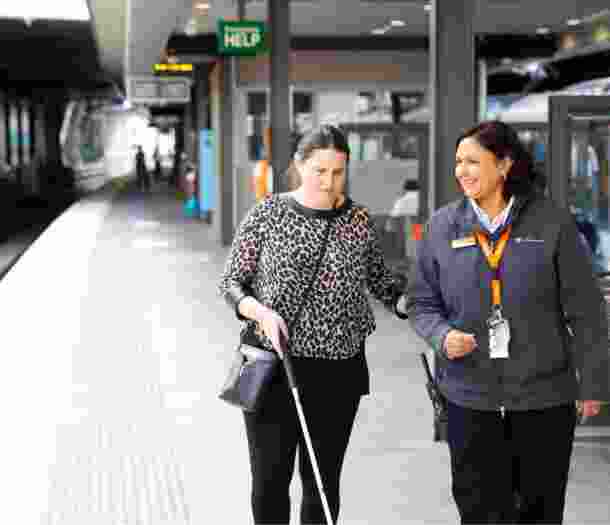 A person using their white cane at the train station while being guided by a train station attendant.