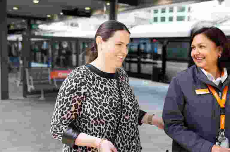A person using their white cane at the train station while being guided by a train station attendant.