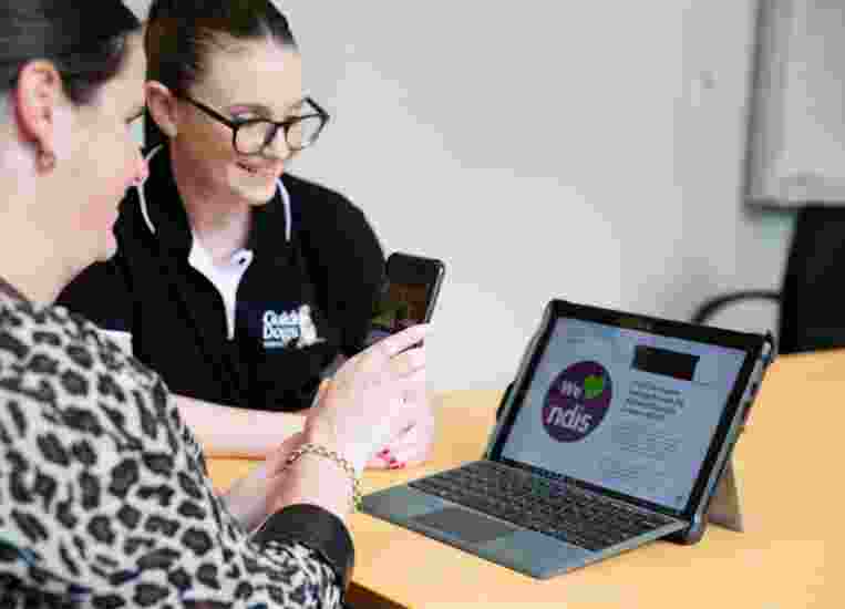 A person and a Guide Dog's staff member looking at a laptop. The person is holding up an Iphone to the laptop and the staff member is smiling.