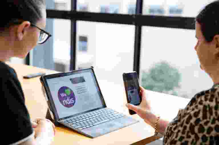 Two people looking at a laptop screen. One is holding a phone to the laptop screen and the other is watching on.