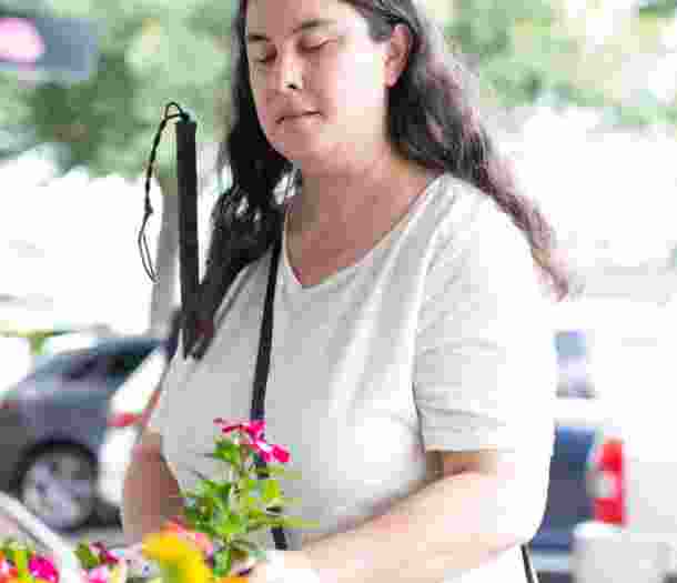 A person is standing in the street holding some flowers in one hand and their white cane in their other arm.