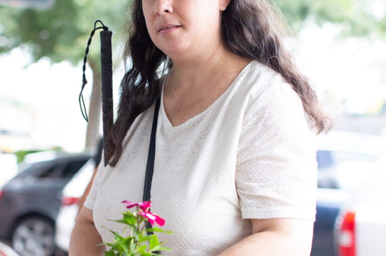 A person is standing in the street holding some flowers in one hand and their white cane in their other arm.