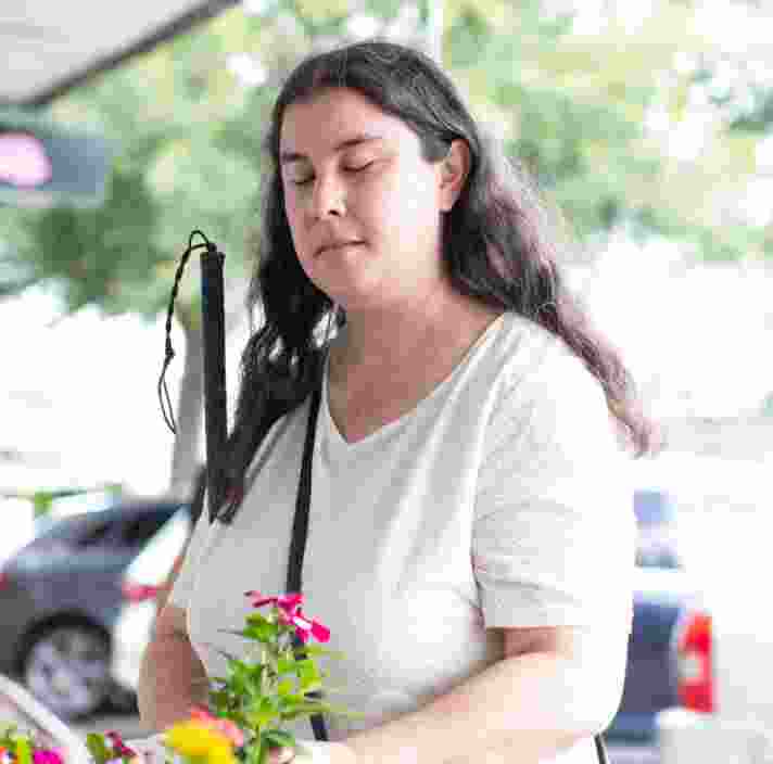 A person is standing in the street holding some flowers in one hand and their white cane in their other arm.