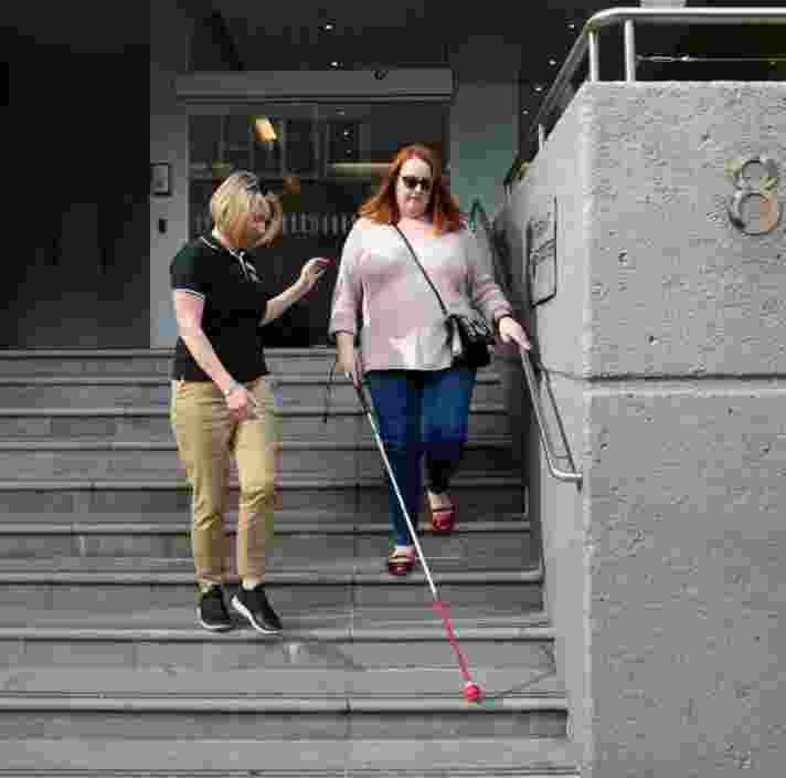 A person assisting a person using a white cane down a set of stairs.