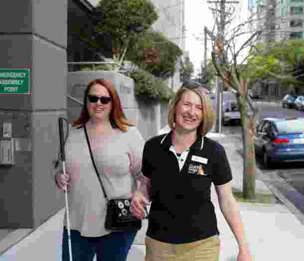 A person with their white cane walking down the street with a Guide Dogs staff member. Both people are smiling at the camera and the person with the cane is holding the staff member by the elbow.