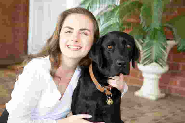 A person looking at the camera smiling with their arm around the neck of a black labrador dog.