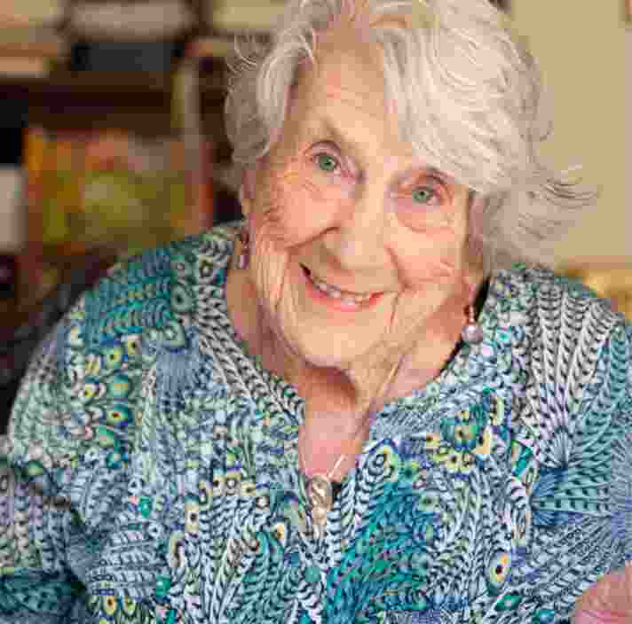 An older adult sitting at a table with a book. The person is looking at the camera smiling.