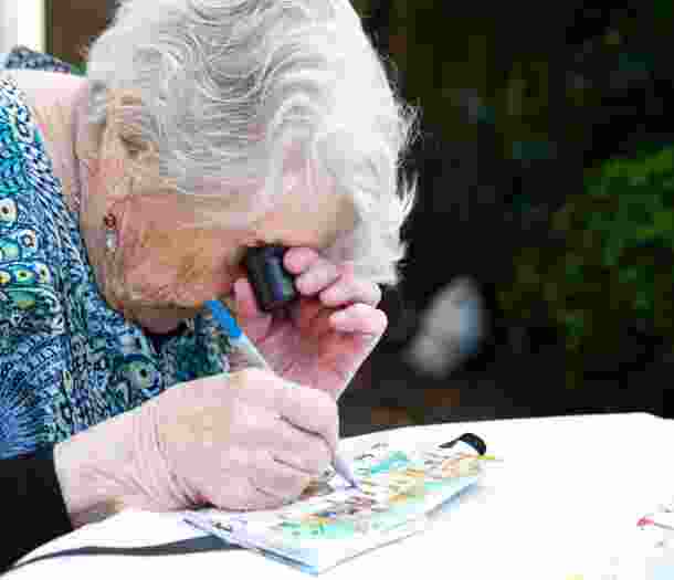 An older adult using a magnifier whilst painting, The person is painting onto a canvas.