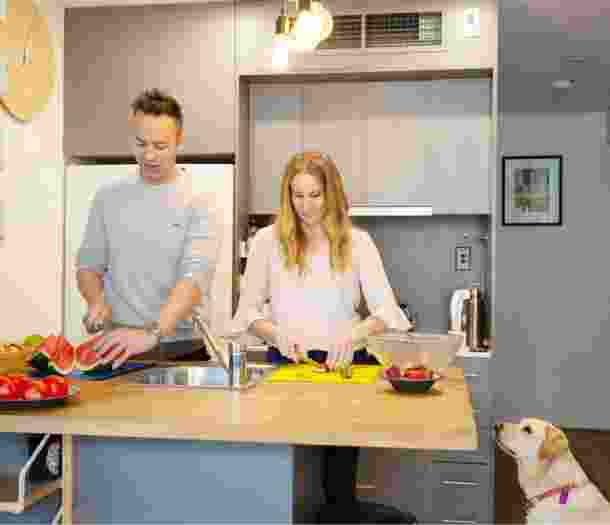 A couple preparing fresh food in their kitchen. Next to the kitchen table is a yellow labrador dog sitting on its back legs looking up at the food.