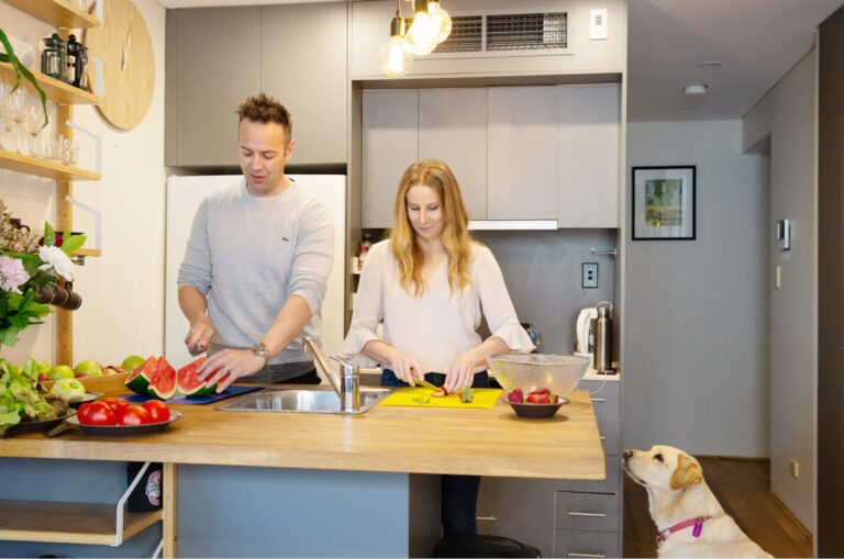 A couple preparing fresh food in their kitchen. Next to the kitchen table is a yellow labrador dog sitting on its back legs looking up at the food.