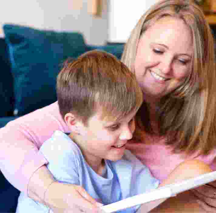 Young child sitting on the ground with their mother next to them. They are both smiling and looking at an Ipad.