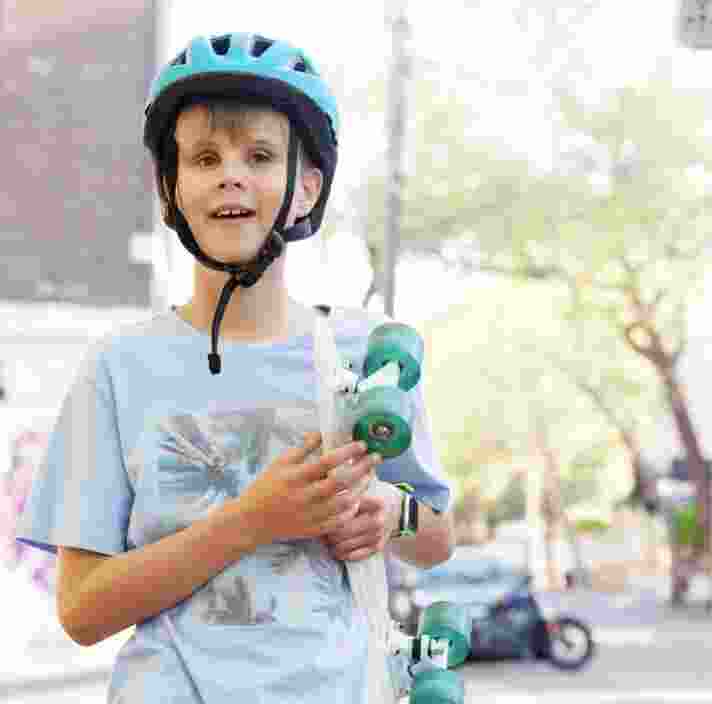 Young child wearing a helmet, holding a skateboard and standing outside in the city streets. The child is looking at the camera and smiling.