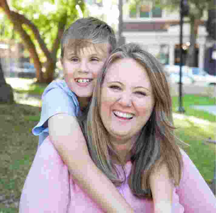 Young child with their arms around their mothers neck and they are both looking at the camera smiling. They are seated on grass outside.