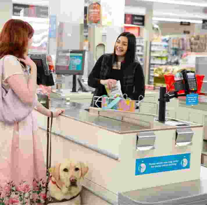 A person at a check out who is looking at the cashier. The cashier is smiling. The person at the checkout has a yellow labrador Guide Dog in harness who is looking at the camera.