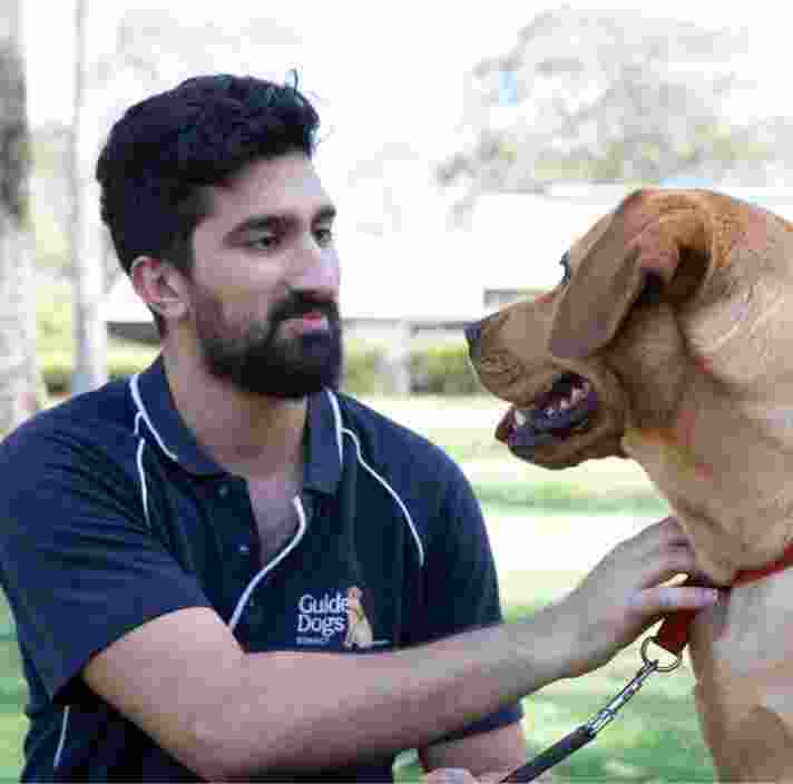 A Guide Dogs staff member and a caramel labrador dog sitting outside. The staff member and dog are both looking at each other.