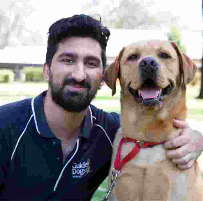 A Guide Dogs staff member and a caramel labrador dog sitting outside. The staff member has their arm around the dog and both are looking at the camera smiling.