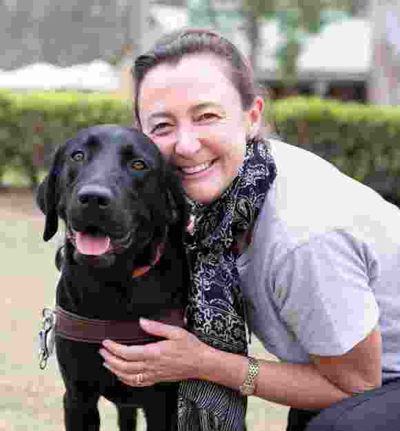 A person hugging a black labrador Guide Dog in harness. The person and dog are looking at the camera and the person is smiling,