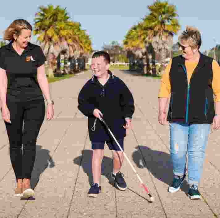 A young boy walking down the path using their cane. On one side of the boy is a Guide Dogs staff member and on the other side is the boys mother. All people have smiles on their faces.