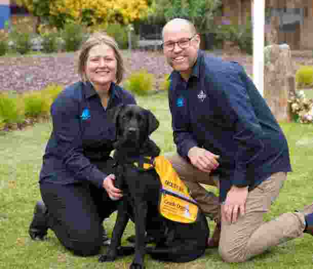 Two people on either side of a black Autism Assistance Dog. The people are kneeling down next to the dog and the photo is taken outside on grass. The dog is wearing an Autism Assistance Dog coat and everyone is looking at the camera.
