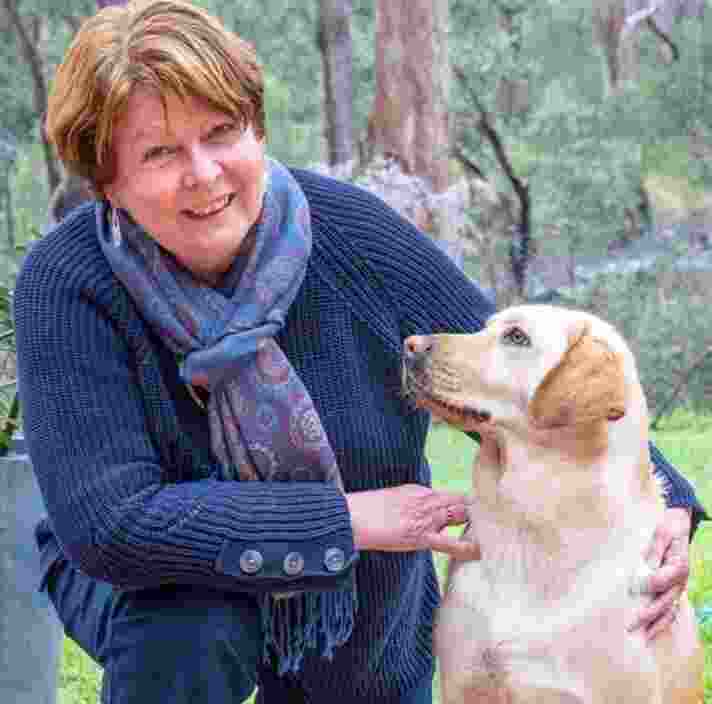 A person kneeling outside next to a yellow labrador dog. The person is looking at the camera smiling and the dog is looking at the person.
