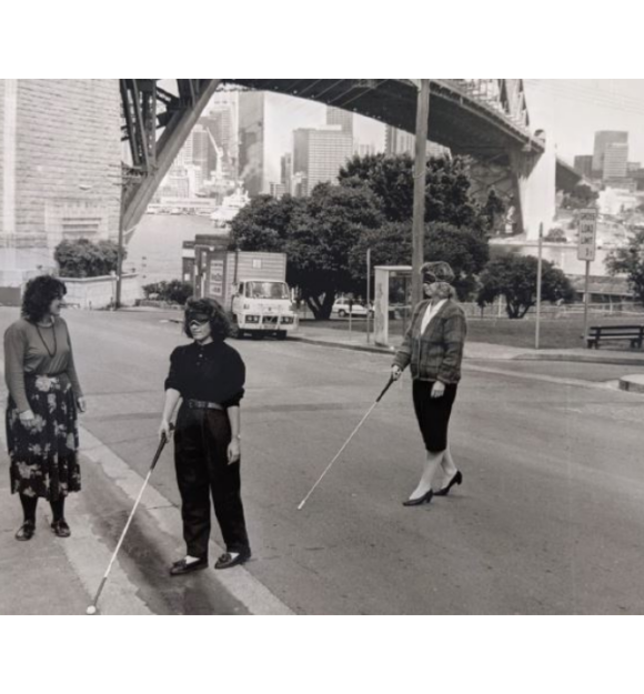A black and white photo of two people blindfolded crossing the street using white canes