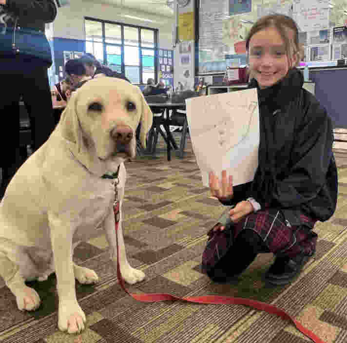 A child holding a drawing, seated next to a yellow Labrador.