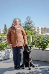 A woman standing next to a black Labrador on a balcony with planter boxes in the background.