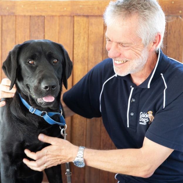 A Guide Dogs staff member and a yellow labrador dog sitting outside. The staff member is patting the dog whilst smiling and they are both looking at each other.