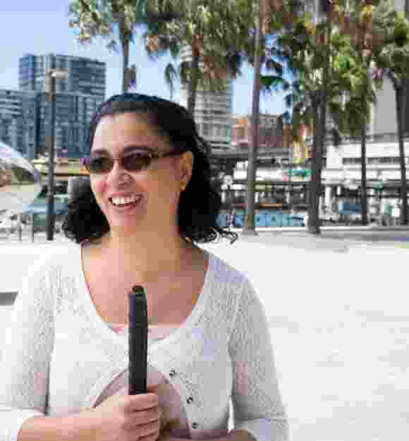 A person standing outside wearing sunglasses and holding a white cane.