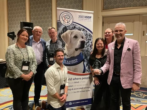 A group of Guide Dogs staff members standing in front of a banner at the IGDF conference.