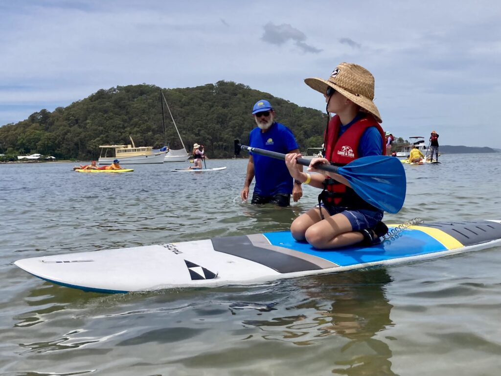 A child kneels on a paddle board holding a blue oar while an instructor stands beside him in the thigh-deep water. 