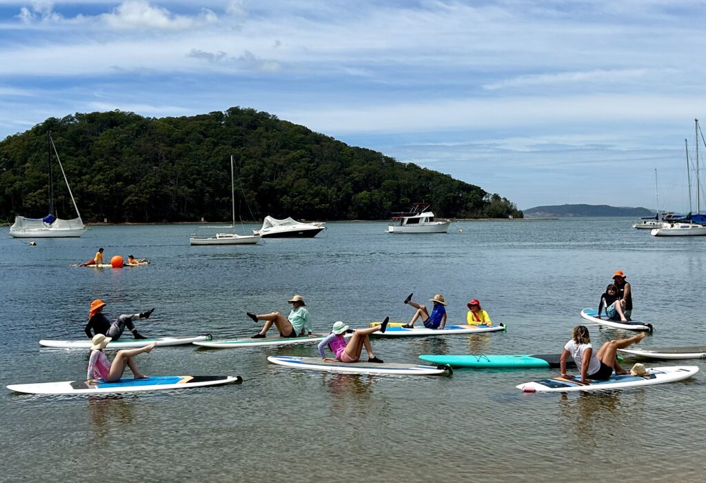 Seven people on paddle boards practice a sitting yoga pose with one leg raised.