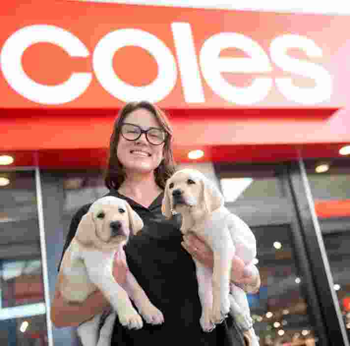 Guide Dog Trainer Chelsea holding two yellow Labrador pups outside a Coles store.
