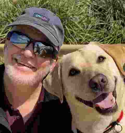 A man and a yellow Labrador Guide Dog look at the camera smiling.