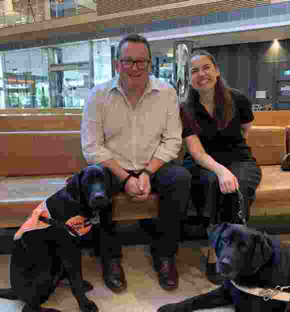 Two people sitting on a bench. At their feet are two black labradors, one wearing an orange training jacket and one in Guide Dog Harness.