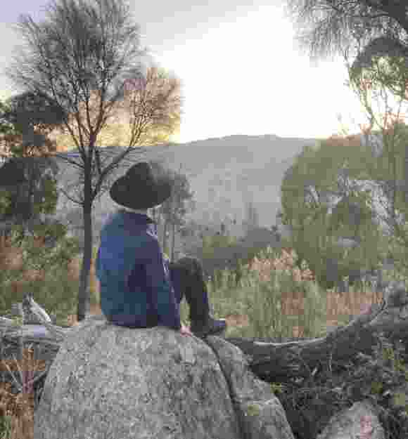 A young man wearing a black wide brim hat sits on a rock in the bush and looks over trees and hills at the sunrise.