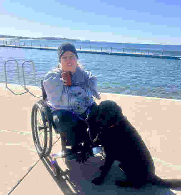 A young woman in a wheelchair and her black Labrador Therapy Dog sit together on a wharf in front of an ocean pool.