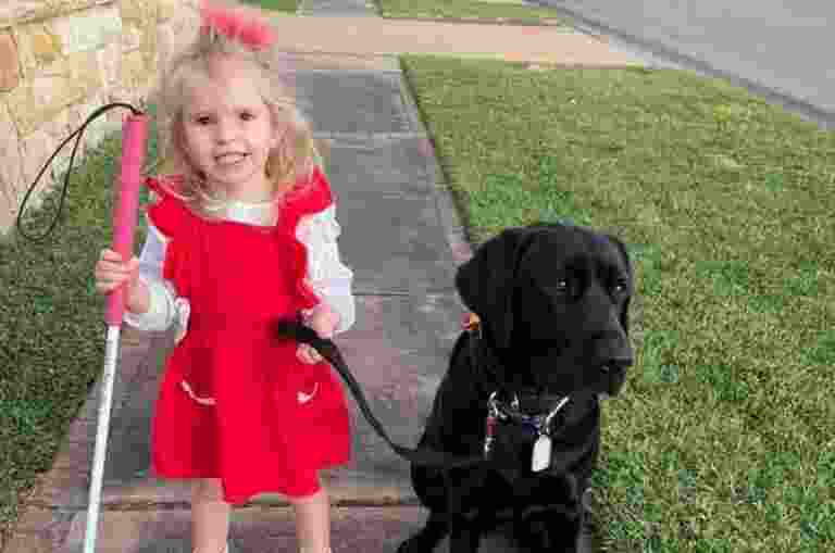 Ivy, a young girl, holds a white cane and stands next to black Labrador Perry outside.