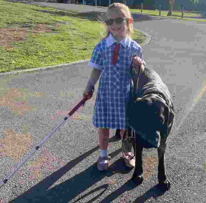 Ivy, a young girl, walking down the street holding on to black Labrador Perry with one hand, and her white cane in the other.