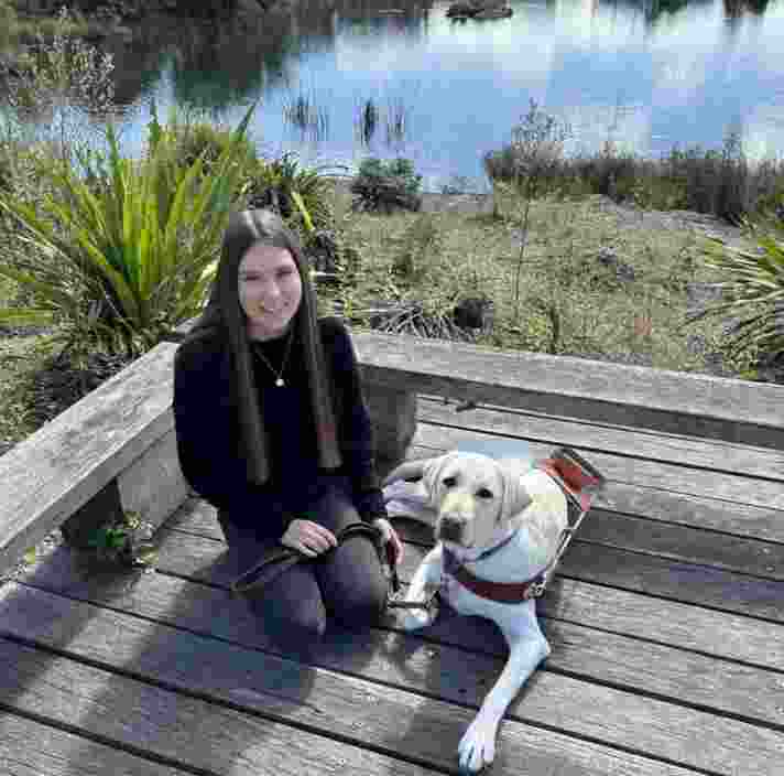 A young woman kneeling next to her yellow Labrador Guide Dog on a wooden boardwalk.