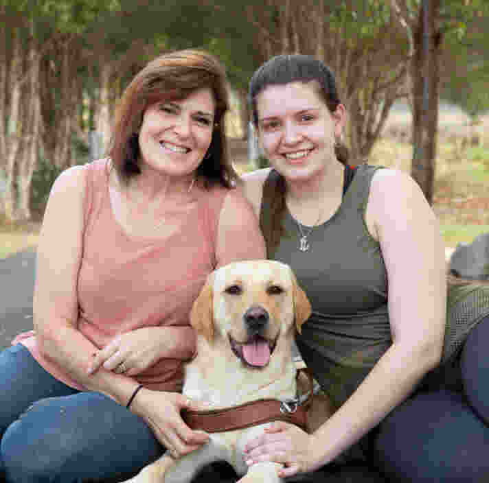 Twp Volunteers sit with a yellow Labrador puppy between them.