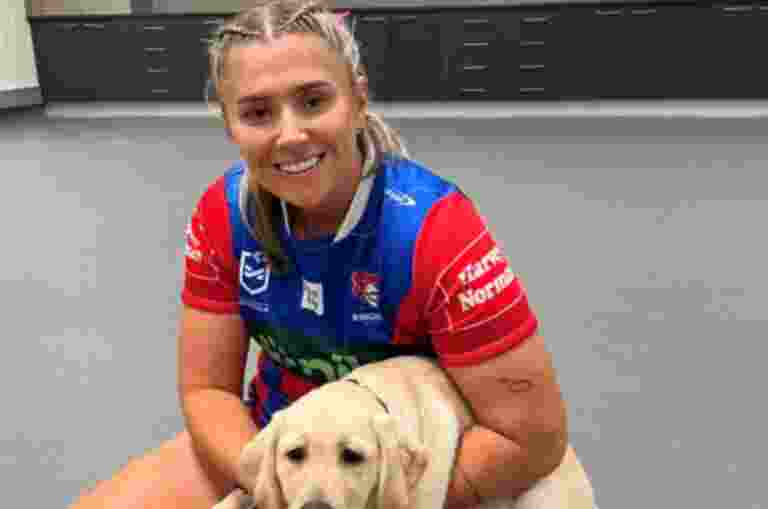 An NRLW Knights team member sitting on the ground holding a yellow Labrador puppy.